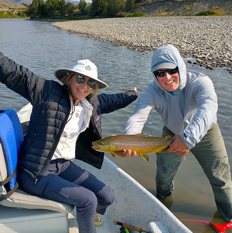 Happy angler and guide with brown trout. Image contains river, rocks, people, and trout.