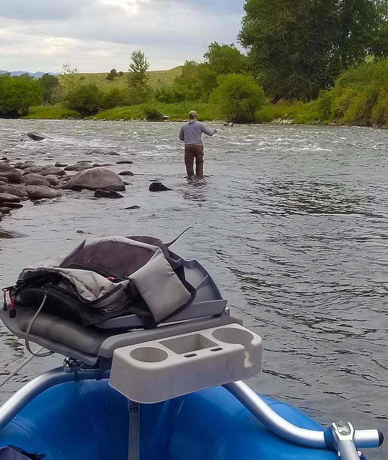 Angler fishing the Boulder River. Photo contains a raft, a river, an angler, and vegetation and sky in background.