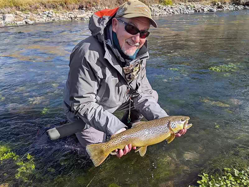 kneeling angler holding large brown trout with river in background and clear water and aquatic plants in foreground